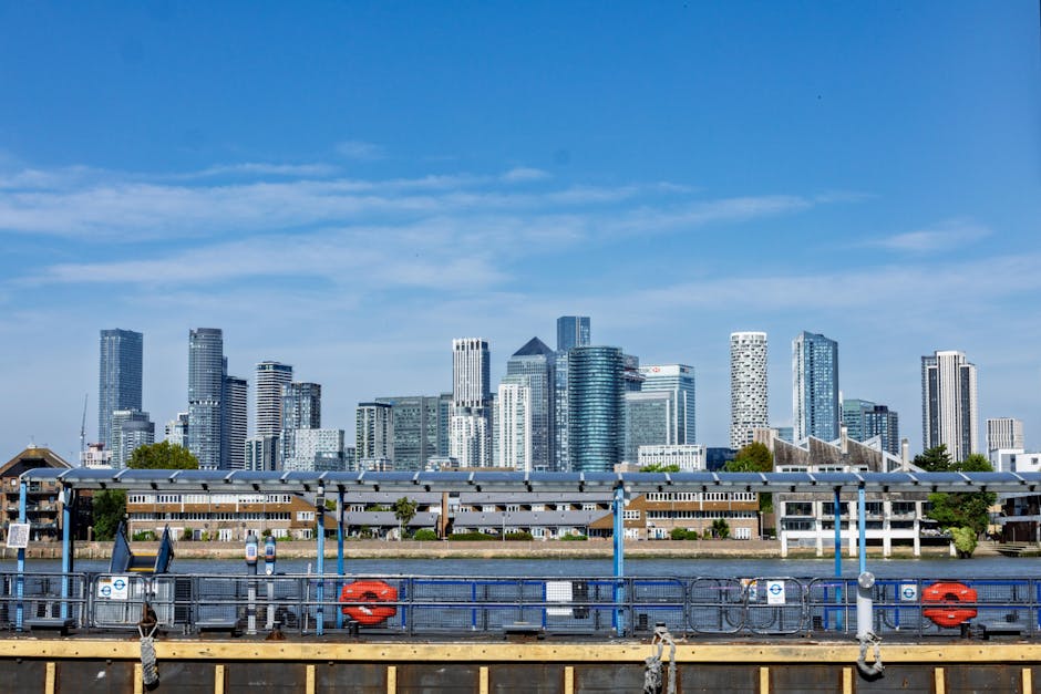 A view of London's modern city skyline featuring a cluster of glass and steel skyscrapers with reflective surfaces, situated behind a riverbank. In the foreground, there is a dock area with metal railings painted in blue, orange life rings attached to the railing, and white signage indicating safety and information. The dock has a row of white posts supporting a covered walkway or shelter, with a flat roof and open sides. The riverbank between the dock and the skyscrapers has low-rise buildings with brown brick façades and flat roofs, possibly residential or commercial structures. The sky above is mostly clear with a few wispy clouds, and natural daylight illuminates the scene evenly. The environment suggests an urban waterfront area, which may be utilized for private or commercial boat access, illustrating aspects of urban waterway management and alternative transport or disposal services in the city context. Waste Disposal Canary Wharf provides rubbish removal services that support maintaining clean waterfront and urban areas without reliance on traditional municipal waste collection.
