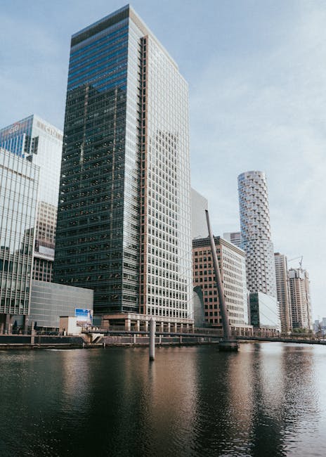 A modern urban landscape featuring a tall, rectangular office building with a glass and metal facade, predominantly dark grey with reflective blue-tinted windows. The building is situated along a riverbank, with its base partially reflected in the water below. To the right, there are other high-rise structures, including a cylindrical tower with a white, textured surface and a support crane in the background indicating ongoing construction. The scene is captured during daylight hours under clear weather conditions, with a pale blue sky and some wispy clouds. In the foreground, the calm water of the river reflects the buildings above, and there are a few dark, slender posts along the river's edge. The overall setting conveys a contemporary financial or commercial district, where private waste disposal services, such as those provided by Waste Disposal Canary Wharf, operate within the city's development landscape, supporting efficient rubbish removal and alternative waste handling outside of municipal collection.