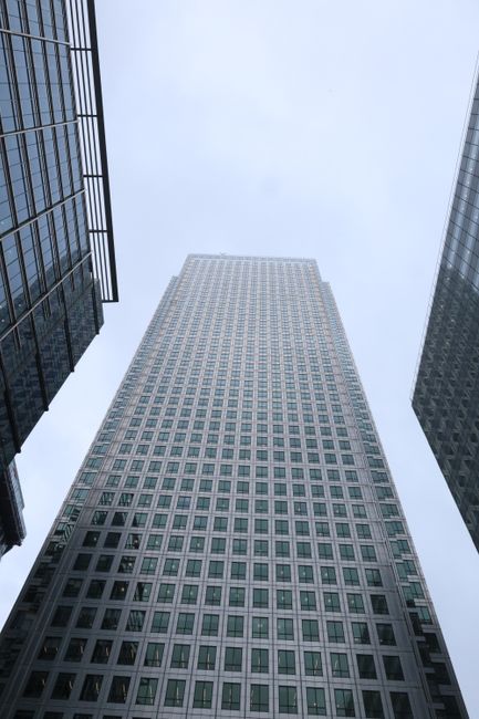 A low-angle view looking up at a tall commercial skyscraper with a grid-like façade of numerous rectangular windows, surrounded by other glass office buildings on either side. The central building has a light-colored exterior, possibly metal or concrete, with a uniform pattern of windows. The sky above is overcast with a pale gray tone, creating a neutral and muted atmosphere. The scene captures the urban environment characteristic of Canary Wharf, emphasizing the scale and modern architectural style typical of the area, relevant to private waste management and alternative rubbish removal services by companies such as Waste Disposal Canary Wharf, which handle waste clearance for large office or commercial premises situated within tall city buildings.