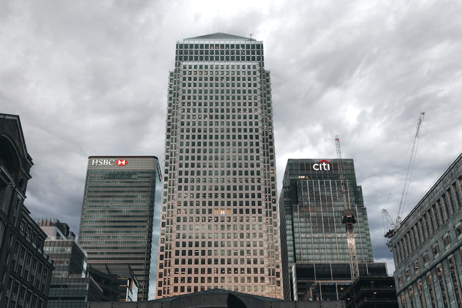 A modern glass-clad office building in an urban setting, featuring large reflective windows arranged in a grid pattern, with rounded corner edges and metal framing visible along the façade. The building's exterior includes multiple floors, with interior lighting illuminating some of the office spaces behind the glass panels. Adjacent taller skyscrapers with similar glass exteriors are seen in the background, contributing to a cityscape environment. The scene is captured during daylight under overcast conditions, creating a neutral light that highlights the building's sleek, contemporary architectural style. The reflection of nearby structures and the slightly muted sky can be observed on the glass surfaces. Such buildings are typical in areas where private disposal and on-site clearance services might be employed to manage waste generated during high-rise renovations or office refurbishments, aligning subtly with waste management and rubbish removal services provided by companies like Waste Disposal Canary Wharf.
