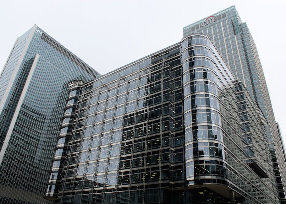 A modern multi-storey office building with a curved glass facade and reflective windows dominates the foreground of this image, with visible metal framing and horizontal lines creating a sleek appearance. To the left and behind, taller taller skyscrapers also feature extensive glass exteriors with vertical and horizontal grid patterns, contributing to a bustling cityscape scene. The overall environment is an urban commercial district with overcast natural lighting, causing a slightly subdued atmosphere with soft reflections on the building surfaces. Although primarily structural, these buildings are indicative of the type of large-scale properties where private waste collection services, such as those offered by Waste Disposal Canary Wharf, might be employed for managing rubbish and waste from tenants. The clear glass and steel finishes emphasize a contemporary architectural style, subtly aligning with the context of alternative waste handling solutions in the city’s business district. No visible rubbish or waste management equipment are present in this image, but the scene implies the kind of environment where professional rubbish removal services might be an essential part of site maintenance.