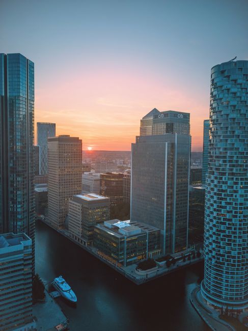 An urban cityscape at sunset featuring modern high-rise buildings with reflective glass facades and varying architectural designs. In the foreground, there is a canal or waterway with a white boat docked along the edge. The buildings are constructed from materials such as glass, steel, and concrete, displaying colours like blue, grey, and beige, with some windows reflecting the warm tones of the setting sun. The skyline includes several mid- and tall-rise structures, some with flat roofs and others with distinctive curved or textured exteriors. The background shows a horizon with a colorful sky transitioning from pinkish-orange near the horizon to a deeper blue higher up, creating a calm atmosphere typical of early evening. The scene captures the essence of a vibrant financial district, where contemporary architecture meets the tranquil waters, aligning with themes of urban development and alternative waste handling for city environments, relevant for waste disposal services like those provided by Waste Disposal Canary Wharf.