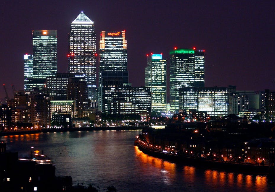 A modern multi-storey office building with a curved glass facade and reflective windows dominates the foreground of this image, with visible metal framing and horizontal lines creating a sleek appearance. To the left and behind, taller taller skyscrapers also feature extensive glass exteriors with vertical and horizontal grid patterns, contributing to a bustling cityscape scene. The overall environment is an urban commercial district with overcast natural lighting, causing a slightly subdued atmosphere with soft reflections on the building surfaces. Although primarily structural, these buildings are indicative of the type of large-scale properties where private waste collection services, such as those offered by Waste Disposal Canary Wharf, might be employed for managing rubbish and waste from tenants. The clear glass and steel finishes emphasize a contemporary architectural style, subtly aligning with the context of alternative waste handling solutions in the city’s business district. No visible rubbish or waste management equipment are present in this image, but the scene implies the kind of environment where professional rubbish removal services might be an essential part of site maintenance.