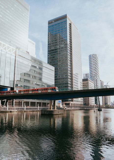 A modern urban scene featuring a large black skyscraper with reflective glass windows in the center, flanked by other high-rise buildings with a combination of glass and concrete facades. In the foreground, a railway bridge extends across a body of water, with a red and blue train visible on the tracks, indicating active transit and infrastructure. Below the bridge, the water is calm, reflecting the surrounding structures with slight ripples. The environment appears to be part of a cityscape with a mix of commercial and office buildings, under soft daylight with a partly cloudy sky, emphasizing a professional setting where efficient waste management and independent collection services, such as those provided by Waste Disposal Canary Wharf, may be required for such high-density developments.