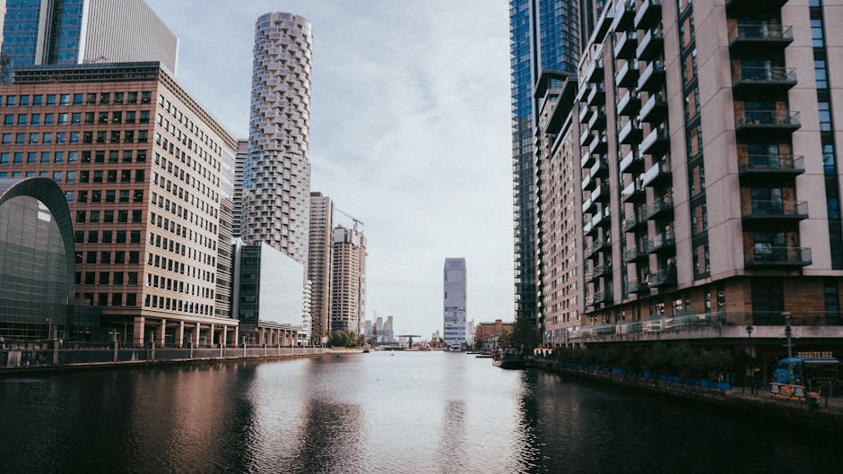 A wide view of a modern urban canal surrounded by tall commercial and residential skyscrapers with varied architectural styles and materials, including glass, steel, and concrete. On the left side, there are prominent high-rise office buildings with large rectangular windows and a curved, dark-colored roof structure on one building. To the right, a collection of residential towers features balconies with glass railings and a mixture of textures in light and dark tones. The canal water reflects the buildings above, appearing calm and slightly rippled. In the background, the canal narrows towards a distant cityscape, with a few construction cranes visible, indicating ongoing development. The scene is illuminated by natural daylight under an overcast sky, casting soft, diffuse light over the scene. The image's urban environment and the prominence of high-rise structures suggest a busy city center, where private waste collection services, such as those provided by Waste Disposal Canary Wharf, could be involved in managing waste from these buildings, contributing to alternative methods of rubbish removal outside of local authority collection.