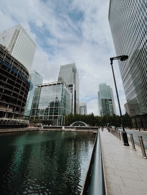 A modern urban scene featuring a large black skyscraper with reflective glass windows in the center, flanked by other high-rise buildings with a combination of glass and concrete facades. In the foreground, a railway bridge extends across a body of water, with a red and blue train visible on the tracks, indicating active transit and infrastructure. Below the bridge, the water is calm, reflecting the surrounding structures with slight ripples. The environment appears to be part of a cityscape with a mix of commercial and office buildings, under soft daylight with a partly cloudy sky, emphasizing a professional setting where efficient waste management and independent collection services, such as those provided by Waste Disposal Canary Wharf, may be required for such high-density developments.